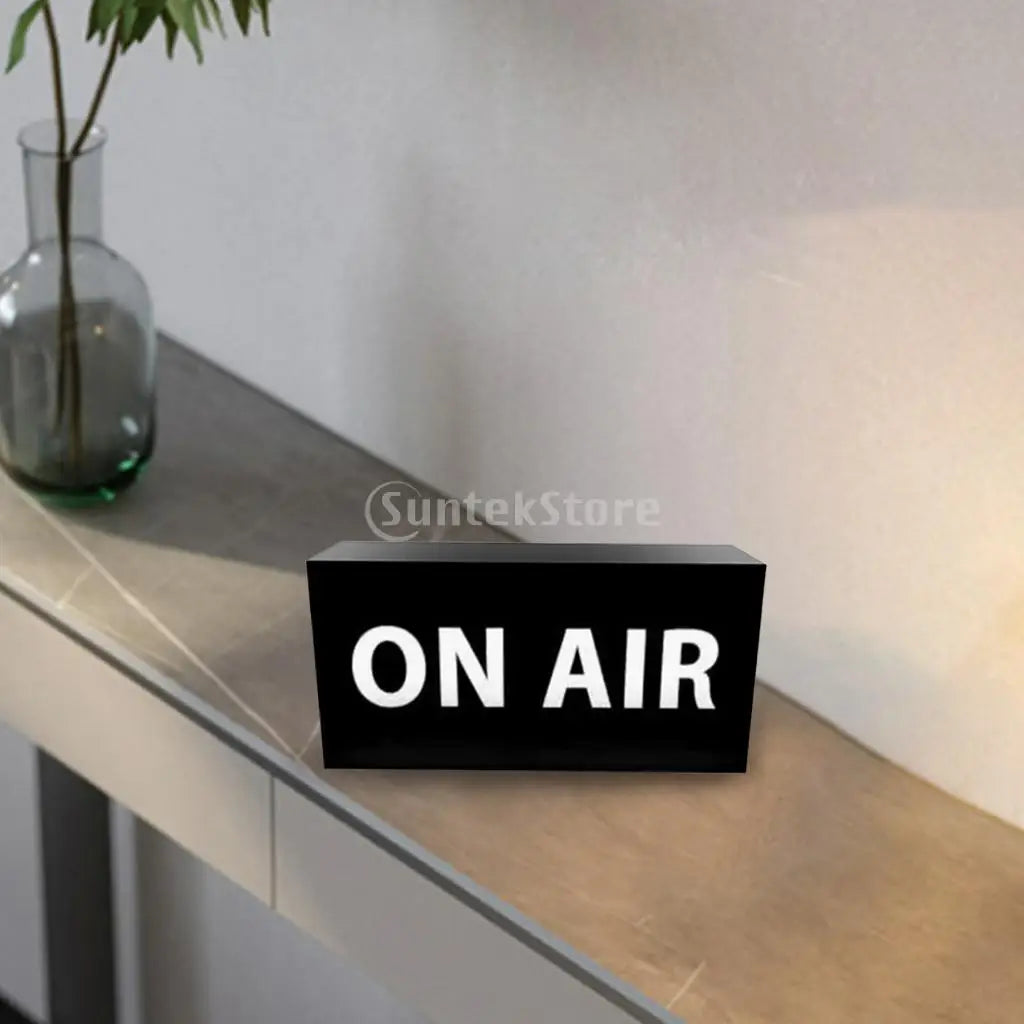 Letrero LED tipo neón “ON AIR” negro con iluminación blanca sobre superficie de mesa.