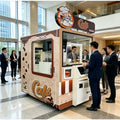 Máquina expendedora robótica de café, cabina beige con letrero “Cafe” y ventana.